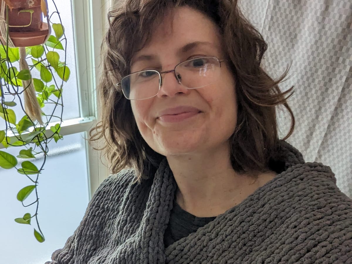 Selfie of a white femme with scrabbly dark hair, gentle face and reading glasses, in a gray knit wrap. Houseplant in bg.