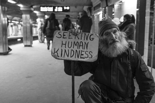BW pic of a man with wild gray beard sitting in a busy subway station holding a sign reading "seeking human kindness."